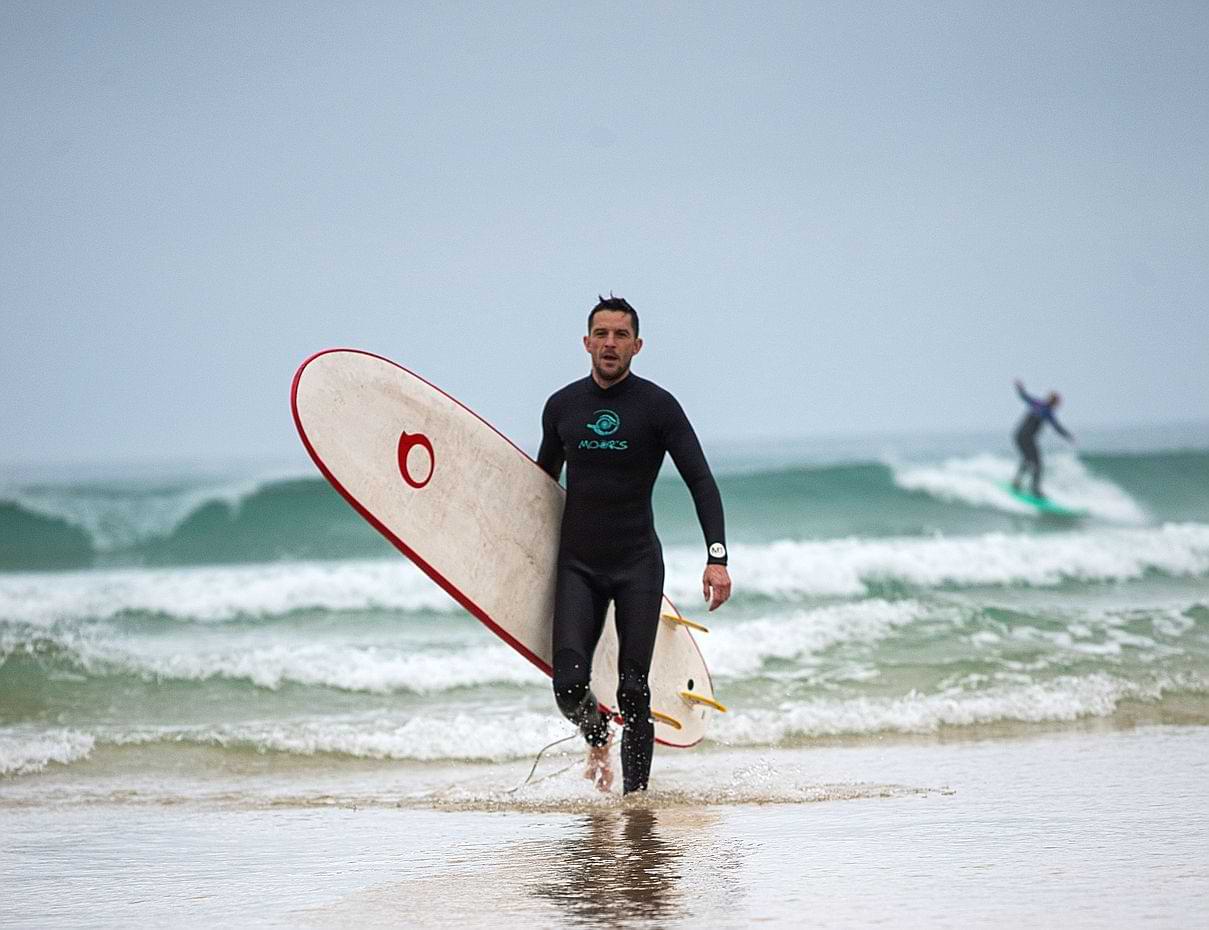 activity Surfer walking on beach with board, another surfing wave
