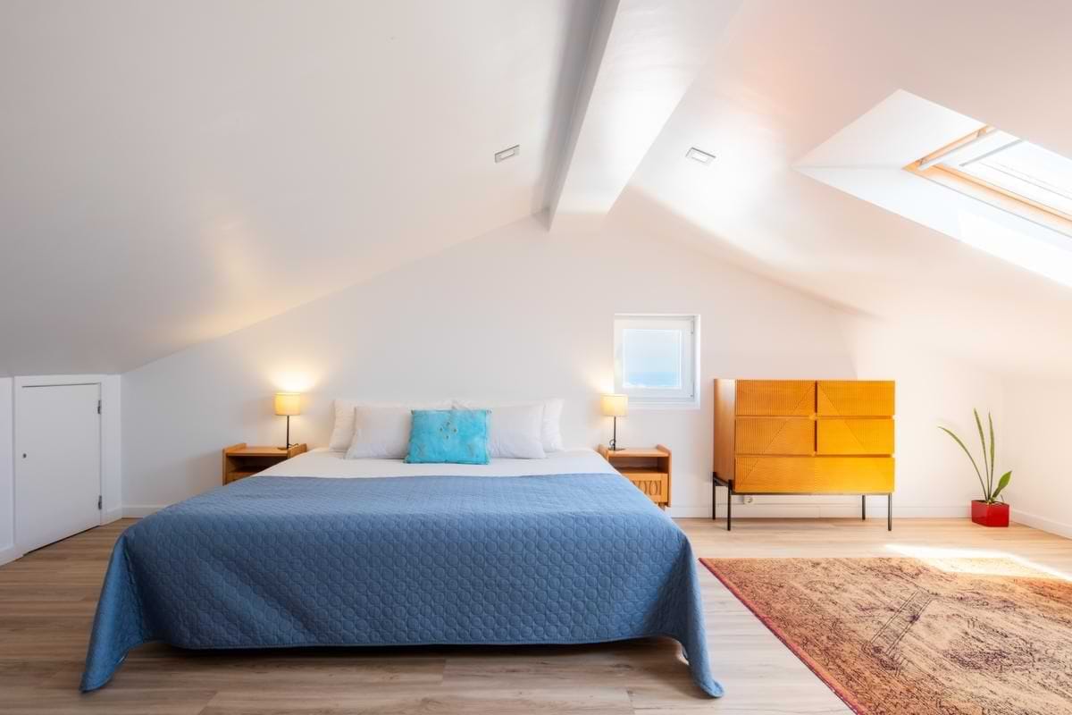 Frontal view of a large double bed centered under a sloped attic ceiling, flanked by bedside tables with lamps, with bright skylights visible to the right.