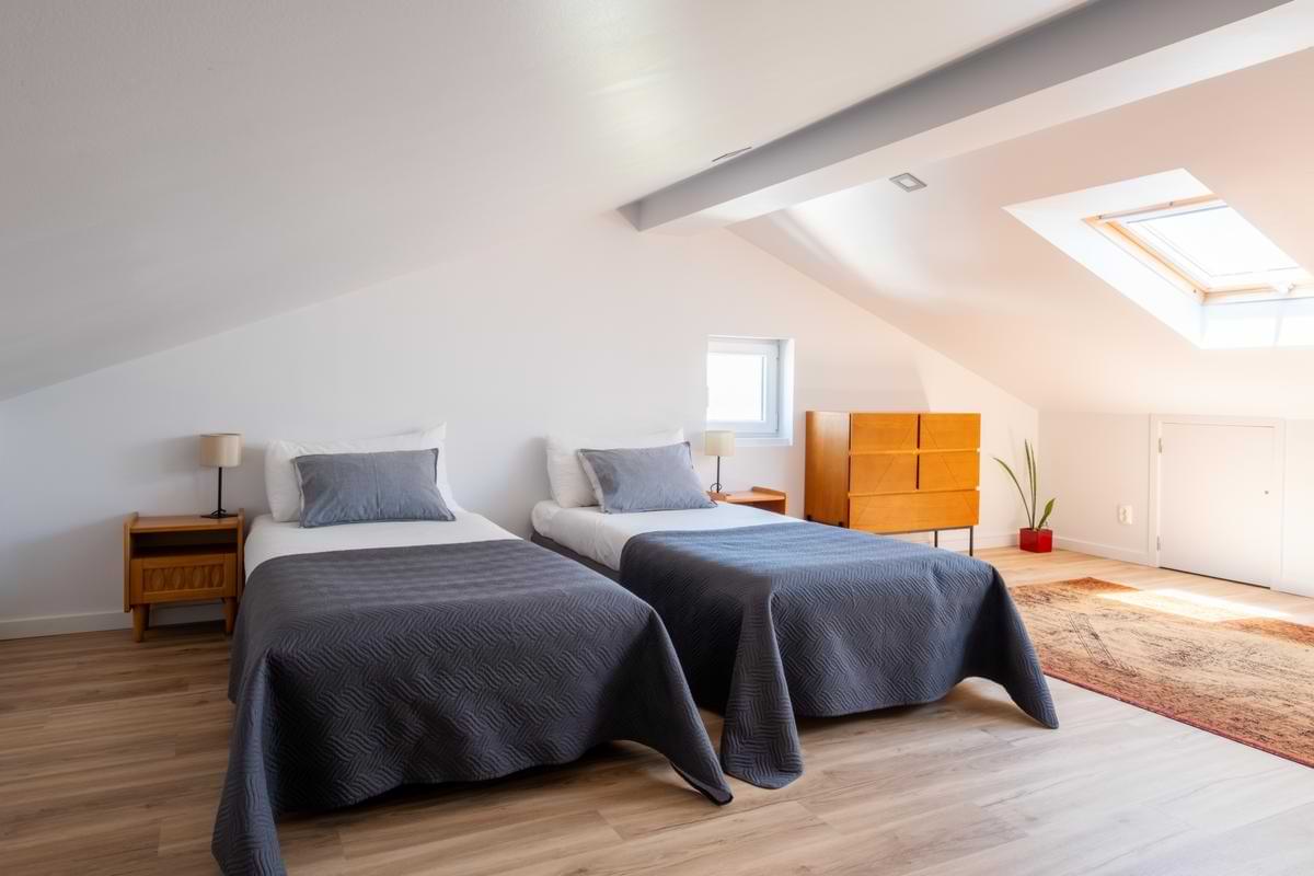 The same attic room configured with two single beds featuring dark grey bedding and grey pillows, positioned under the sloped white ceiling with the orange dresser and rug visible in the background.