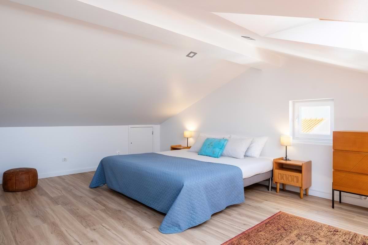 An angled perspective of the attic bedroom showing the length of the double bed with the blue coverlet, a leather pouf in the corner, and a small side window next to a wooden dresser.