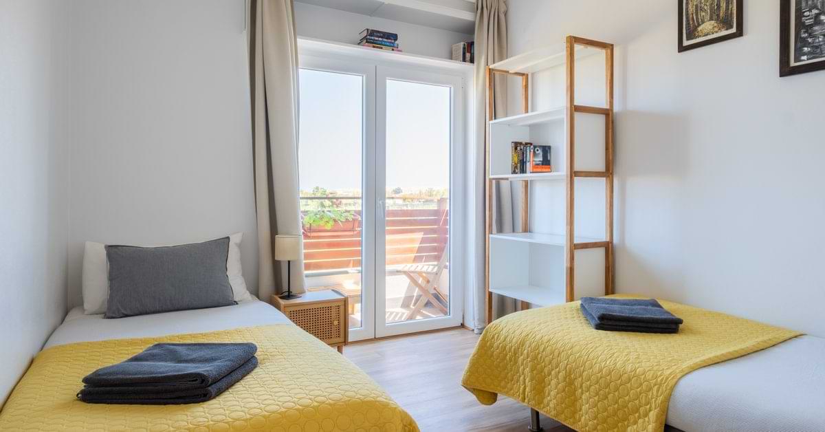 Bright twin bedroom at Surf Atlantic Baleal surf house featuring yellow duvets, gray pillows, and a private balcony door.
