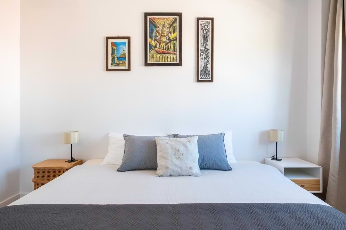 Close-up view of the wall above a double bed, showcasing a curated arrangement of three unique framed pictures, flanked by modern white nightstands with simple lamps.