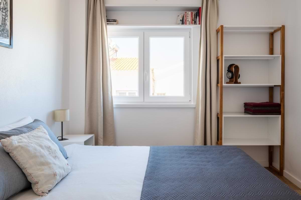 Side view of a bedroom showing a small white desk positioned under a window with beige curtains, a simple chair, and a shelving unit with books and towels.