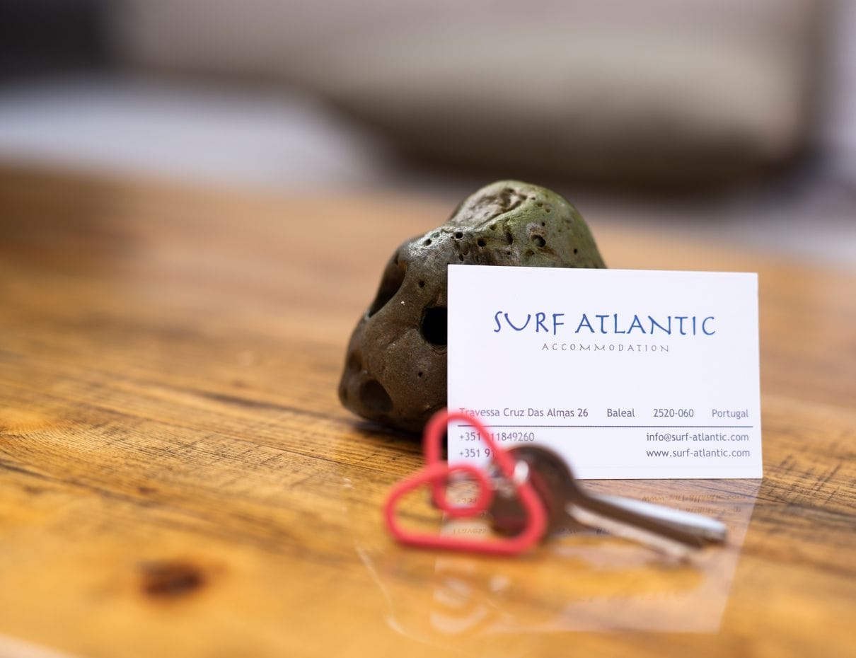 Book Your Surf Holiday House at Surf Atlantic Baleal Surf Atlantic business card with contact details and house keys resting on a wooden table in Baleal, Portugal
