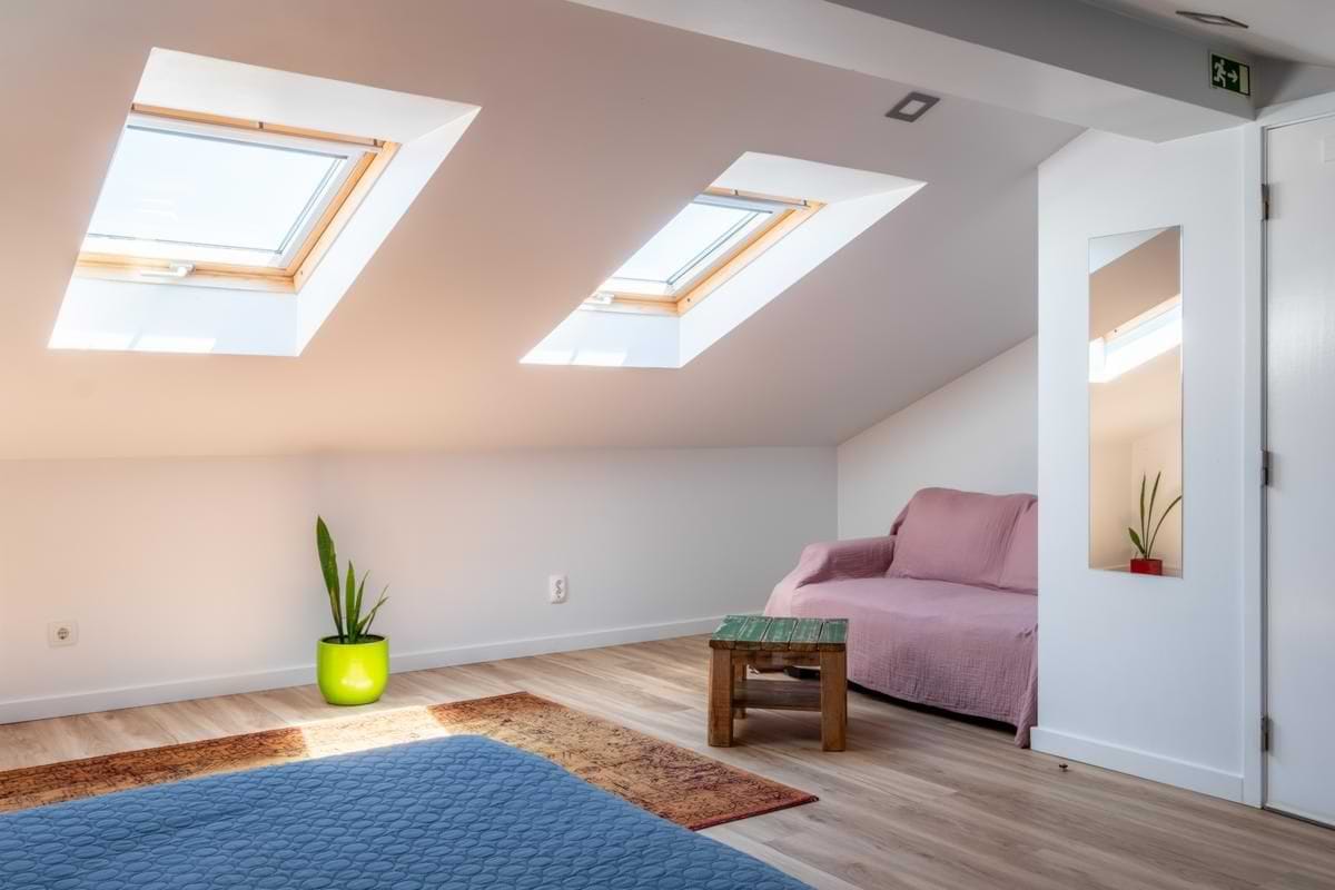 nterior view looking up at two large wooden-framed skylights built into a white sloped attic ceiling, with a pink sofa bed and a lime green plant pot below in the sunbeam