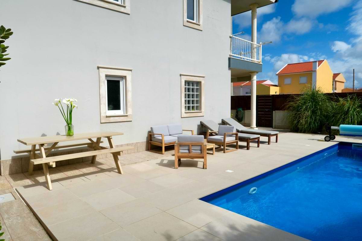 Diagonal wide view of Chillax Villa showing the blue private swimming pool, concrete patio deck, sun loungers, and outdoor dining furniture under a bright sky.