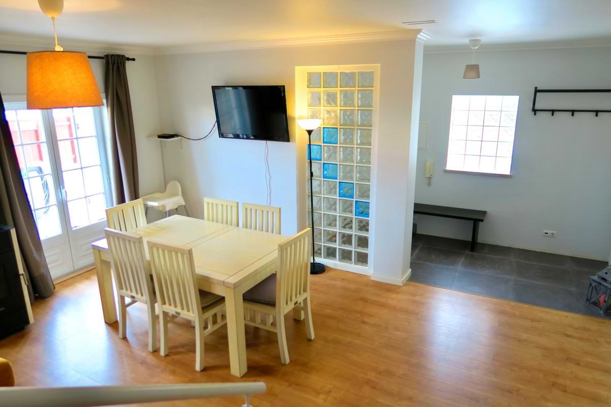 White wooden indoor dining table set for six guests next to a glass-brick accent wall and wall-mounted TV at Chillax Villa.