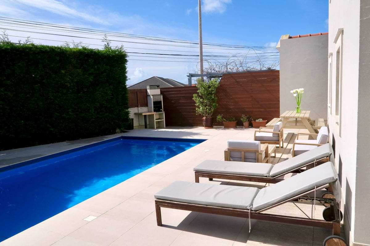 Sun loungers lined up along the private swimming pool next to a stone BBQ grill and garden wall at Chillax Villa.
