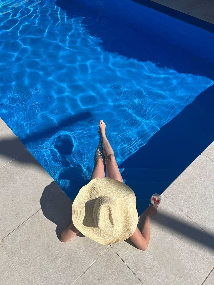 High-angle view of a guest wearing a straw hat sitting on the edge of the blue private swimming pool at Chillax Villa near Peniche.