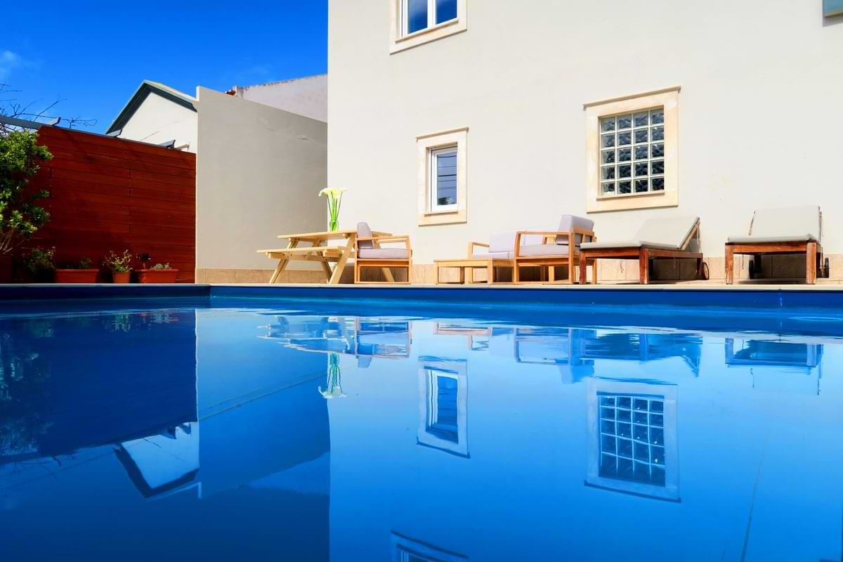 Low-angle wide view showing the house facade and outdoor dining table cleanly reflected in the blue water of the private pool at Chillax Villa