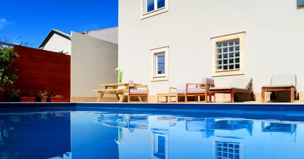A serene pool with clear blue water reflecting a white house, surrounded by a garden and patio furniture.