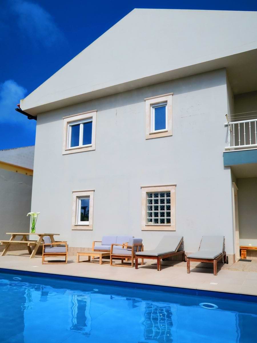 A modern house with a pool in front, featuring lounge chairs and outdoor seating on a patio. The house has several windows and a light-colored exterior under a bright blue sky.