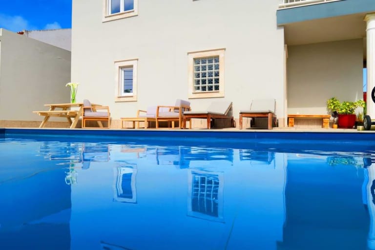 A swimming pool with a clear blue surface reflecting the surrounding white house and outdoor furniture.
