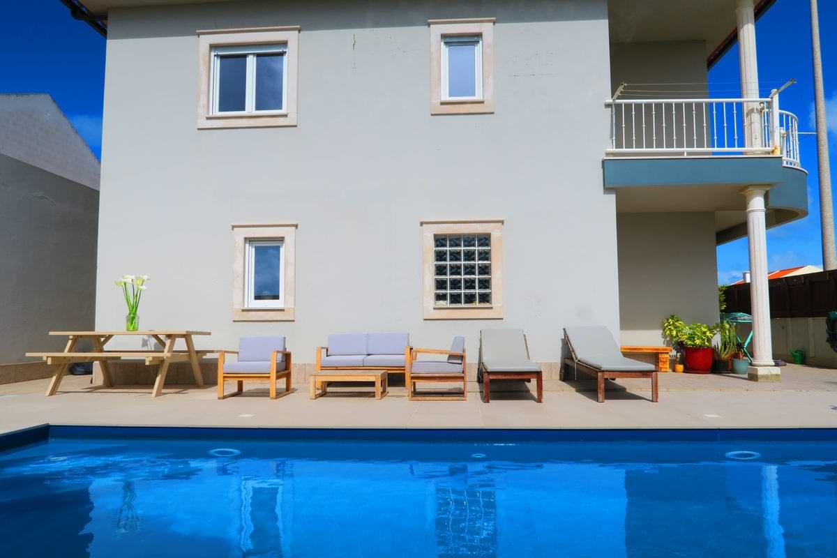 A modern house adjacent to a swimming pool, featuring outdoor seating arrangements, including wooden chairs and loungers, with a table and decorative plants.
