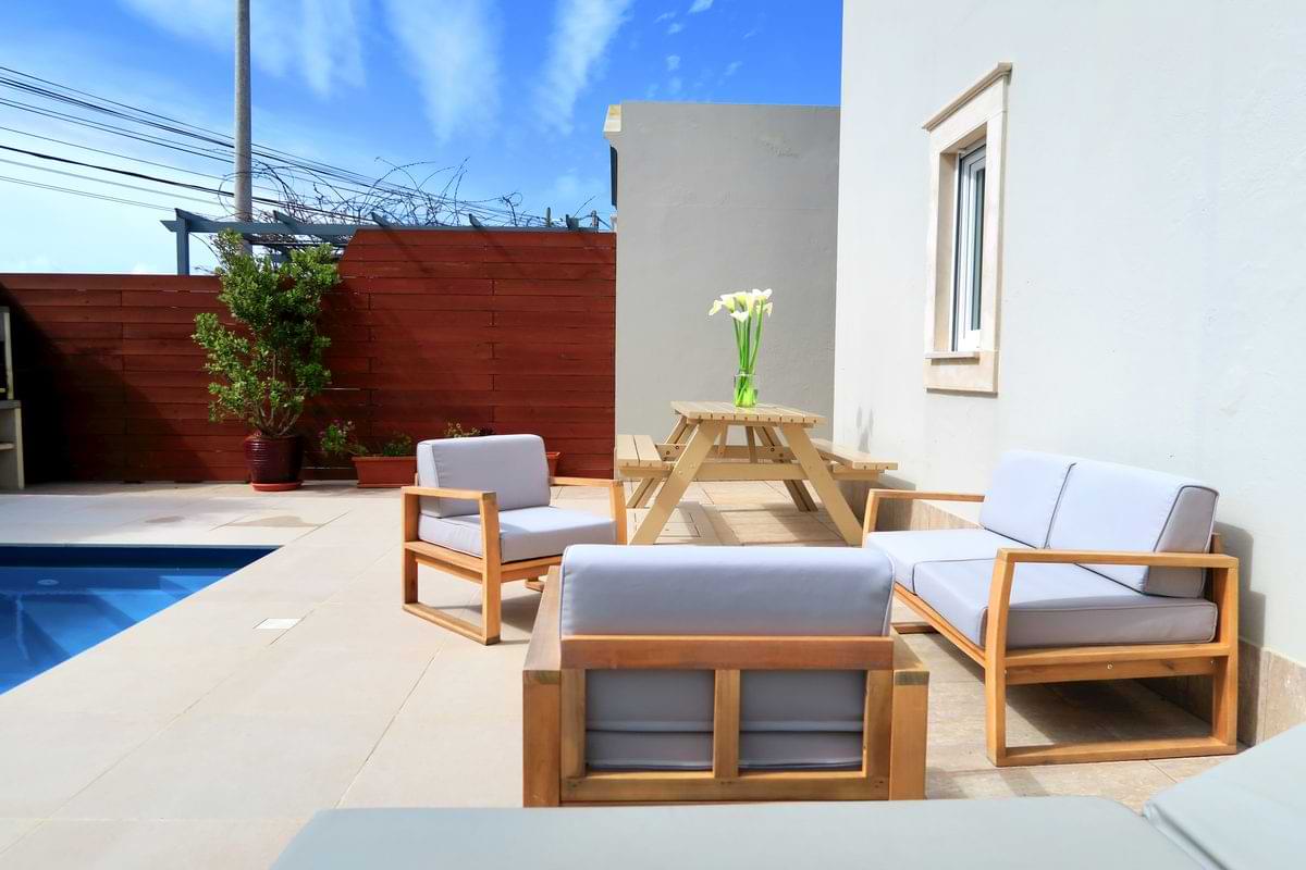 A modern outdoor seating area featuring light grey sofas with wooden frames, a wooden picnic table, and a vase of white flowers on the table. There is a swimming pool nearby and some greenery in the background.