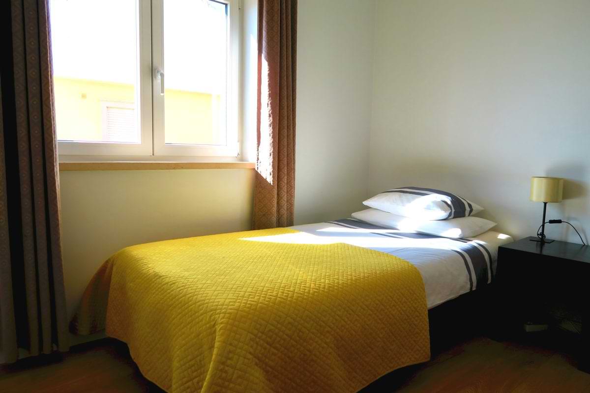 A bedroom with a single bed covered in a yellow blanket, natural light coming through the window, and a lamp on a nightstand.