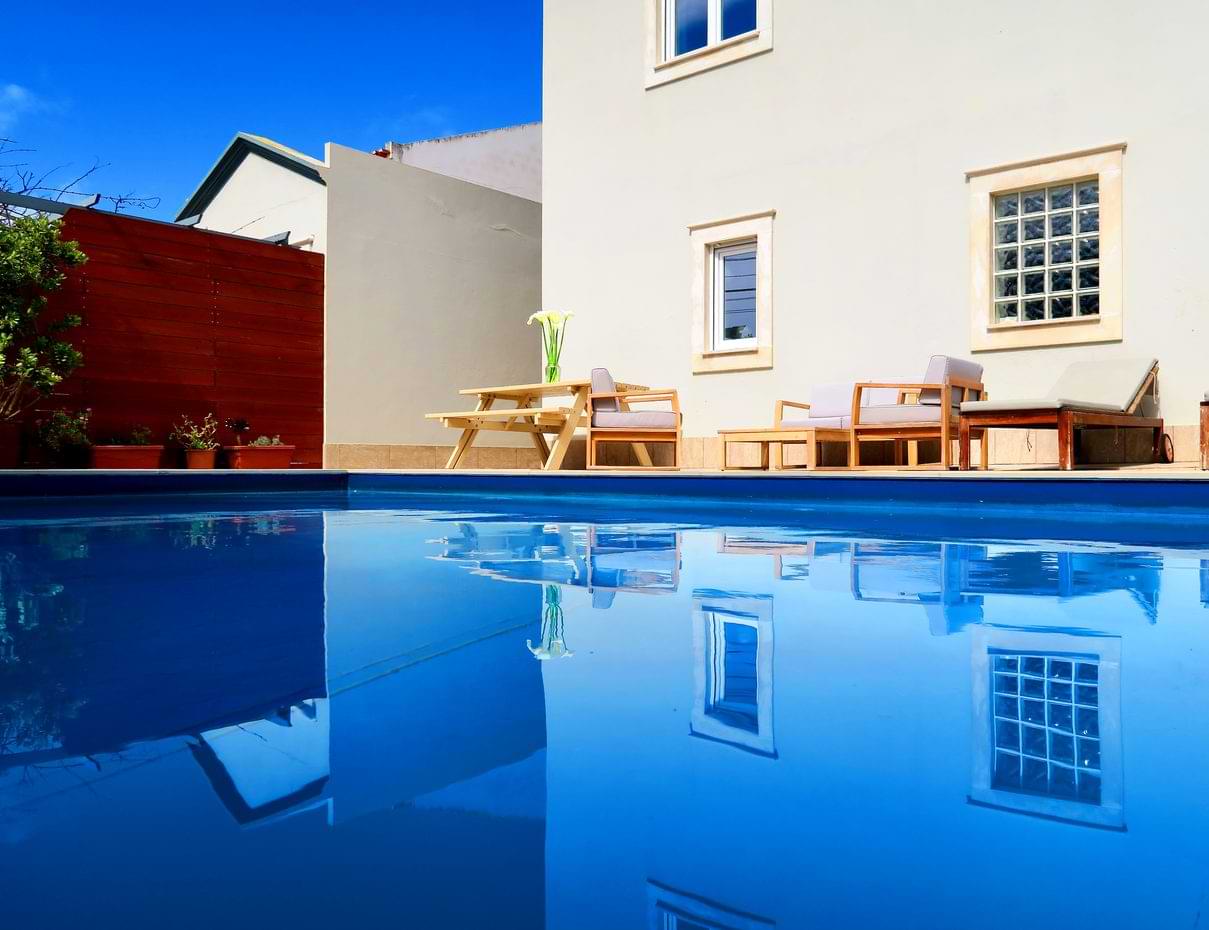 A clear blue pool reflecting a house and furniture in the background, with greenery on one side under a bright blue sky.