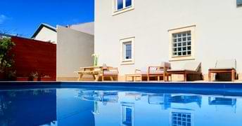 A clear blue swimming pool reflecting a house on a sunny day, with outdoor furniture nearby.