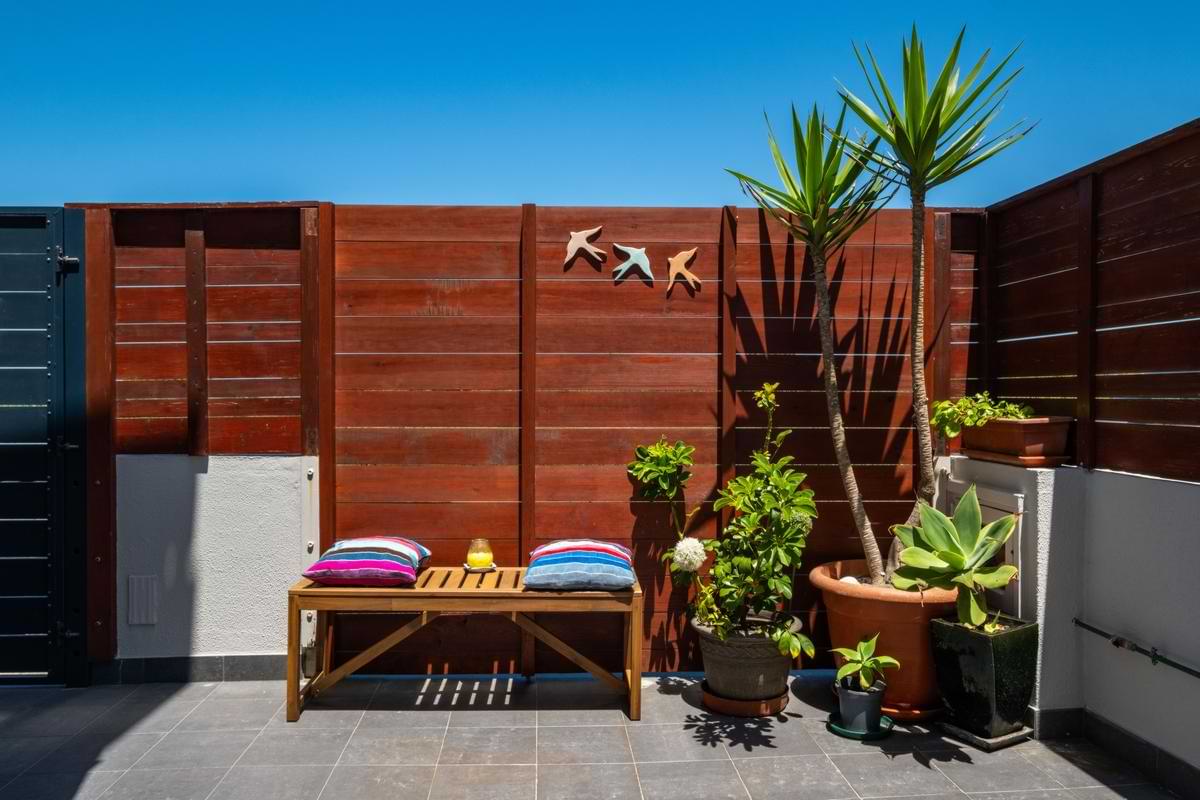 Wooden outdoor bench with colorful cushions positioned against a dark wood fence decorated with metal bird sculptures and lush potted plants.