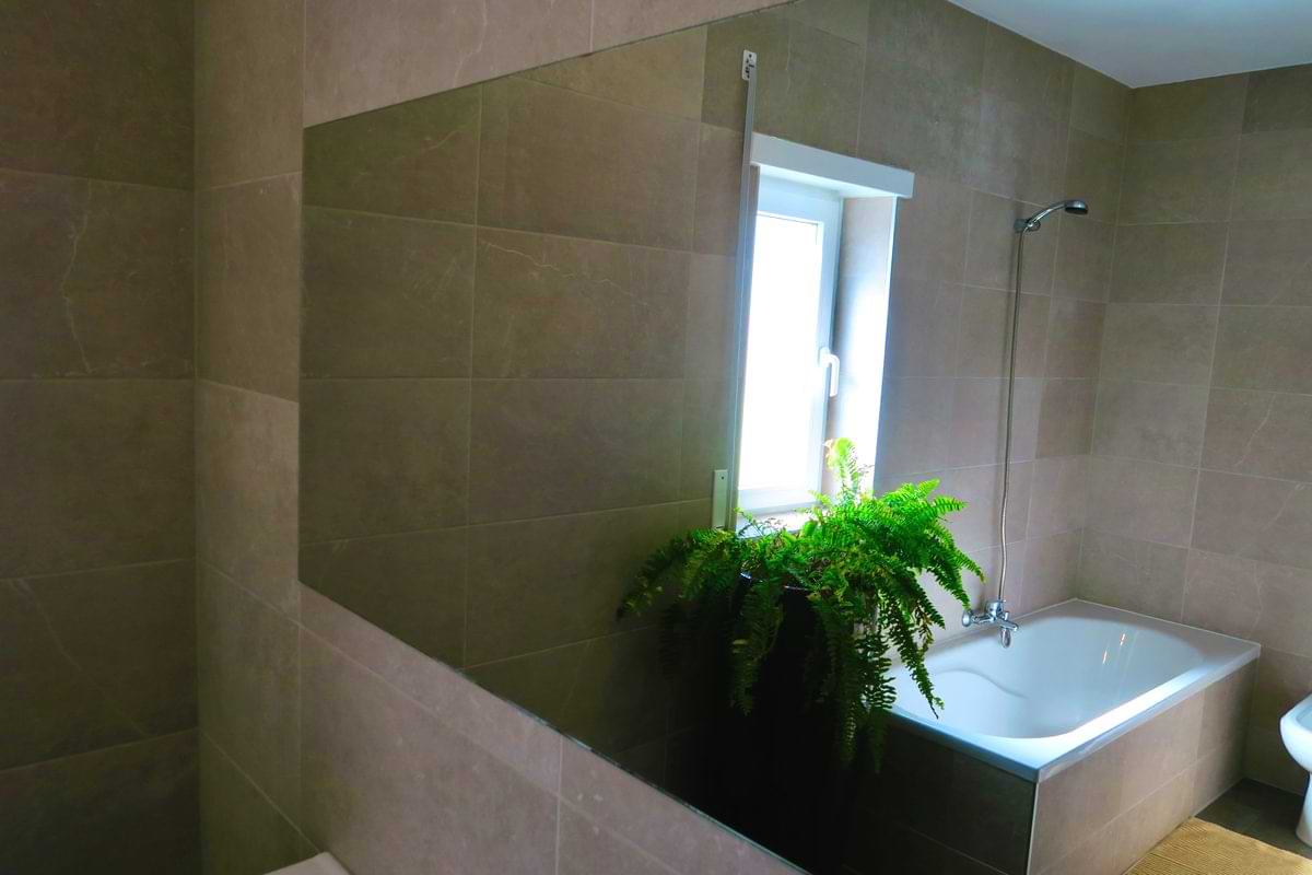 A modern bathroom featuring a bathtub, green potted plant, and a window with natural light. The walls are tiled in neutral tones.