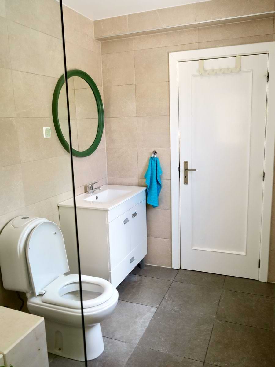 A light-colored bathroom featuring a toilet, a white sink cabinet with a mirror above it, and a blue towel hanging on the wall. There is also a door leading out of the bathroom.