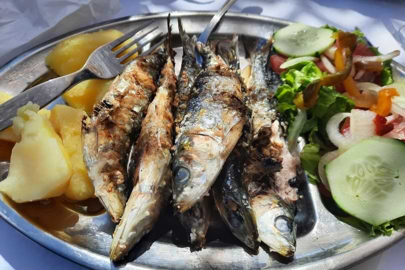 Freshly grilled sardines served on a metal platter with boiled potatoes and a fresh cucumber and tomato salad.