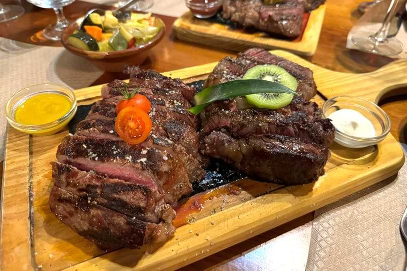 Sliced grilled steak served on a wooden board with cherry tomatoes, kiwi garnish, dipping sauces, and roasted vegetables.