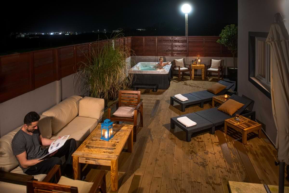 Cozy nighttime view of the illuminated wooden deck with guests relaxing on outdoor sofas and soaking in the hot tub under atmospheric lighting.