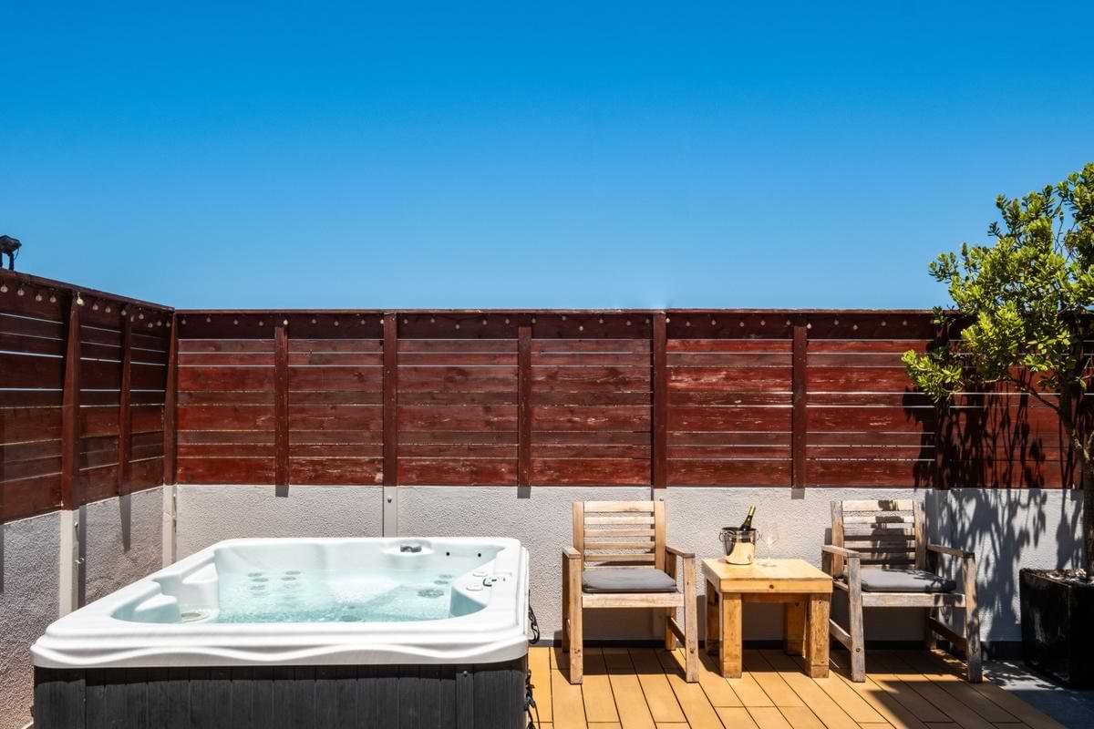 Close-up of the outdoor hot tub alongside two wooden armchairs, a wooden side table with champagne and glasses, set against a dark wood privacy fence.