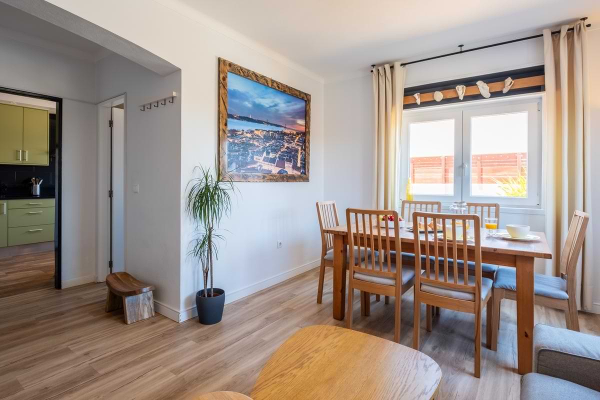 Wooden dining table set for six under a bright window with neutral curtains, featuring a large framed wall art print and a house plant nearby.