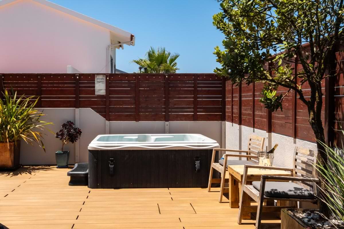 Ground-level view of the hot tub on a composite wooden deck, surrounded by a wooden privacy wall, potted plants, and wooden lounge seating.
