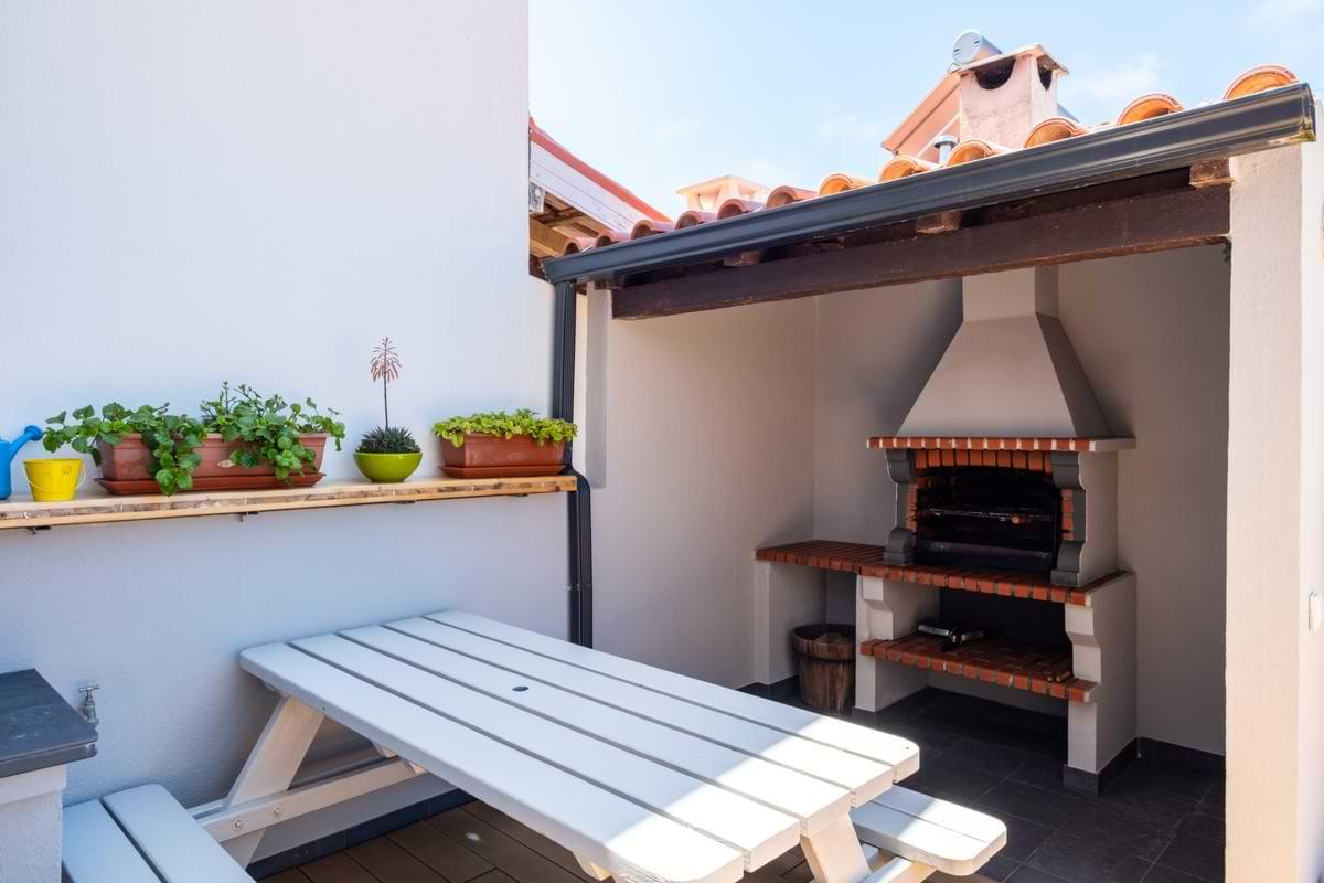Covered outdoor patio nook featuring a traditional stone and brick built-in barbecue grill, a white wooden picnic table, and potted wall plants.
