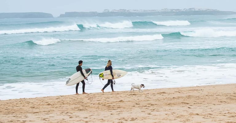 Two surfers in wetsuits carrying surfboards walk along an uncrowded sandy beach in Portugal with a dog.