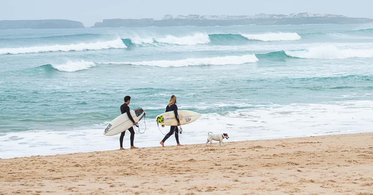 Two surfers in wetsuits carrying surfboards walk along an uncrowded sandy beach in Portugal with a dog.