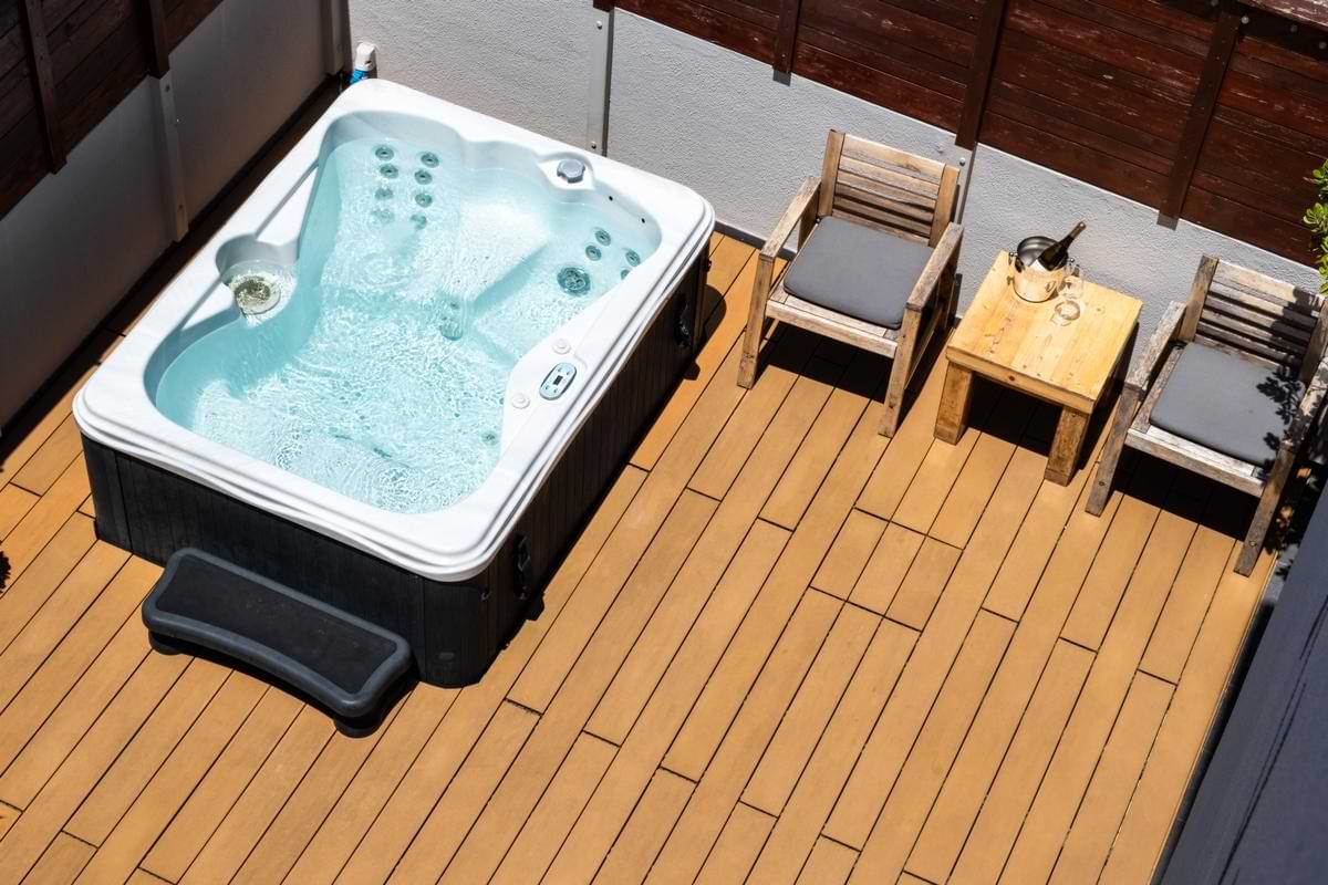 High-angle shot of a hot tub filled with clear bubbling water on a wooden deck, complete with wooden armchairs and a wine bucket on a side table.