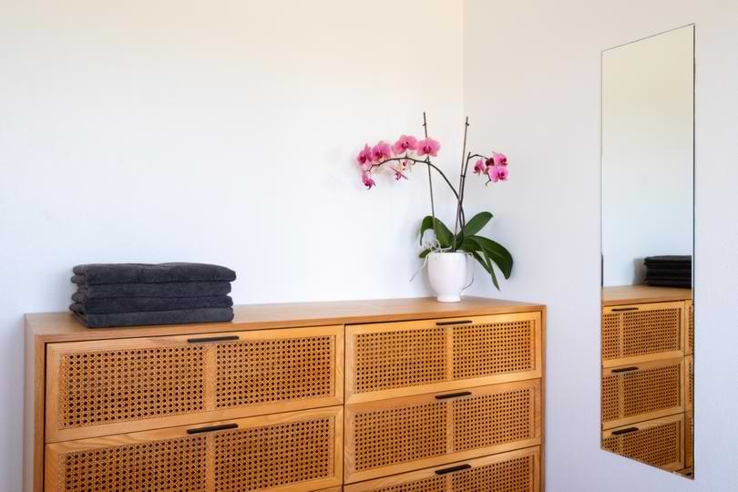 Rattan Bedroom Dresser & Mirror Decor | Surf Atlantic Natural rattan wooden dresser topped with folded towels, a potted pink orchid, and a full-length wall mirror in a Surf Atlantic bedroom.