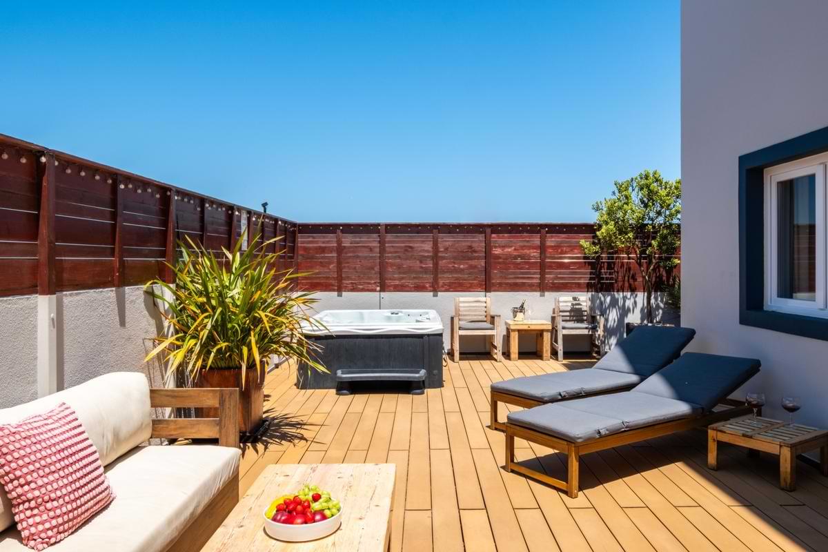 Bright, sun-drenched outdoor patio deck with grey-padded sun loungers, a private hot tub, wooden sofa, and fruit bowl on the coffee table under a clear sky.
