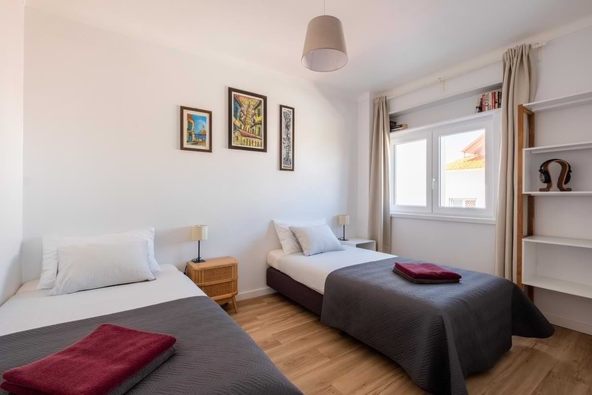 Shared Twin Room Accommodation – Surf Atlantic Baleal Shared twin room featuring two single beds with gray covers, warm wooden flooring, and decorative wall art at Surf Atlantic.
