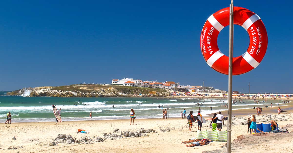 Sunny view of Baleal beach in Portugal with surfers, beachgoers, and a red lifebuoy, capturing a Surf Atlantic guest vacation day.