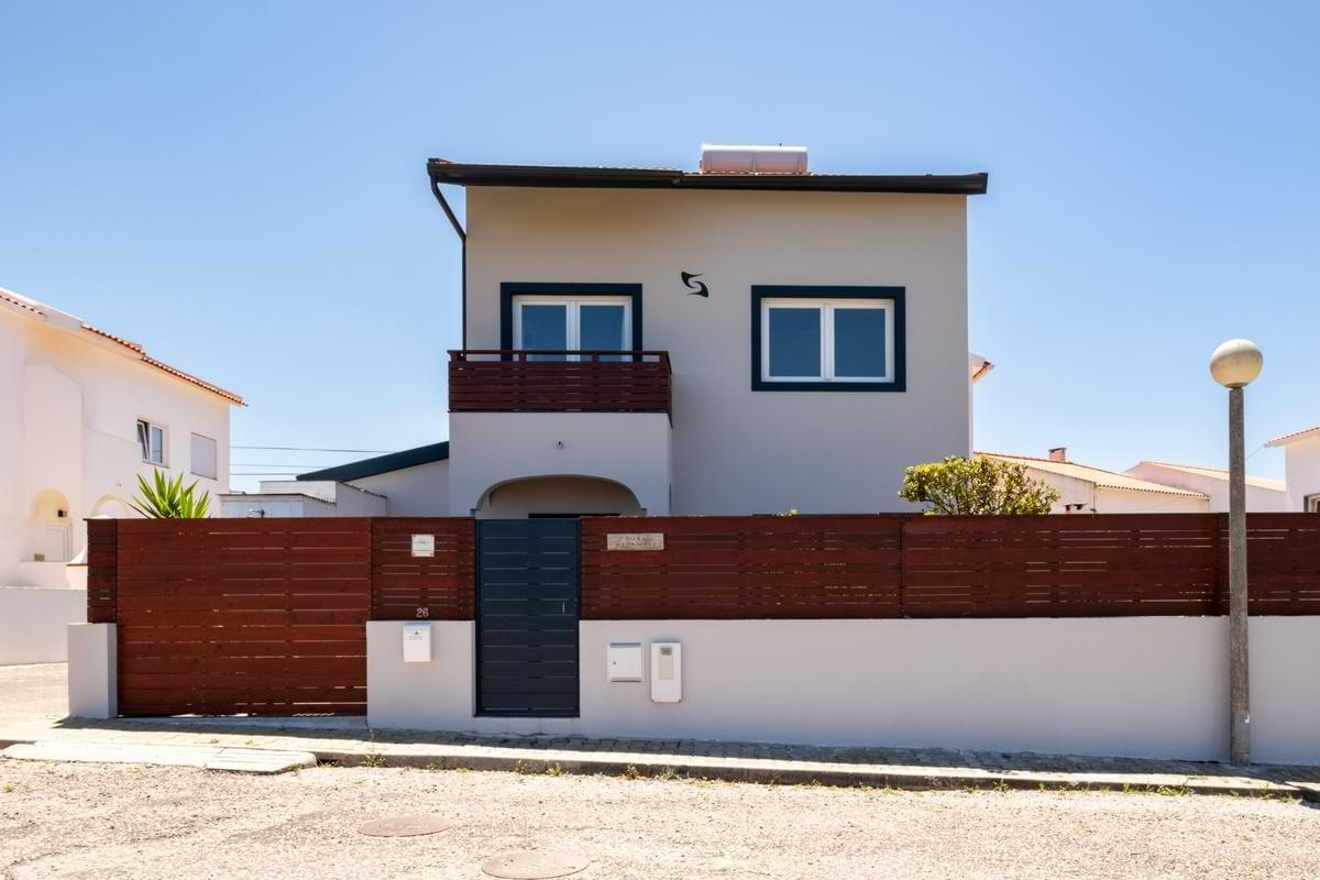 Front view of the two-story Surf Atlantic house featuring white exterior walls, dark blue window trims, a dark grey privacy gate, and a wooden fence.