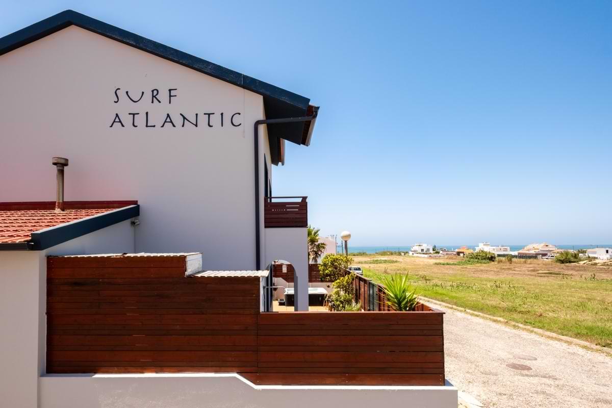 Side elevation of the Surf Atlantic house displaying the wall logo, modern wooden fence perimeter, and distant view of the Atlantic Ocean under a clear blue sky.