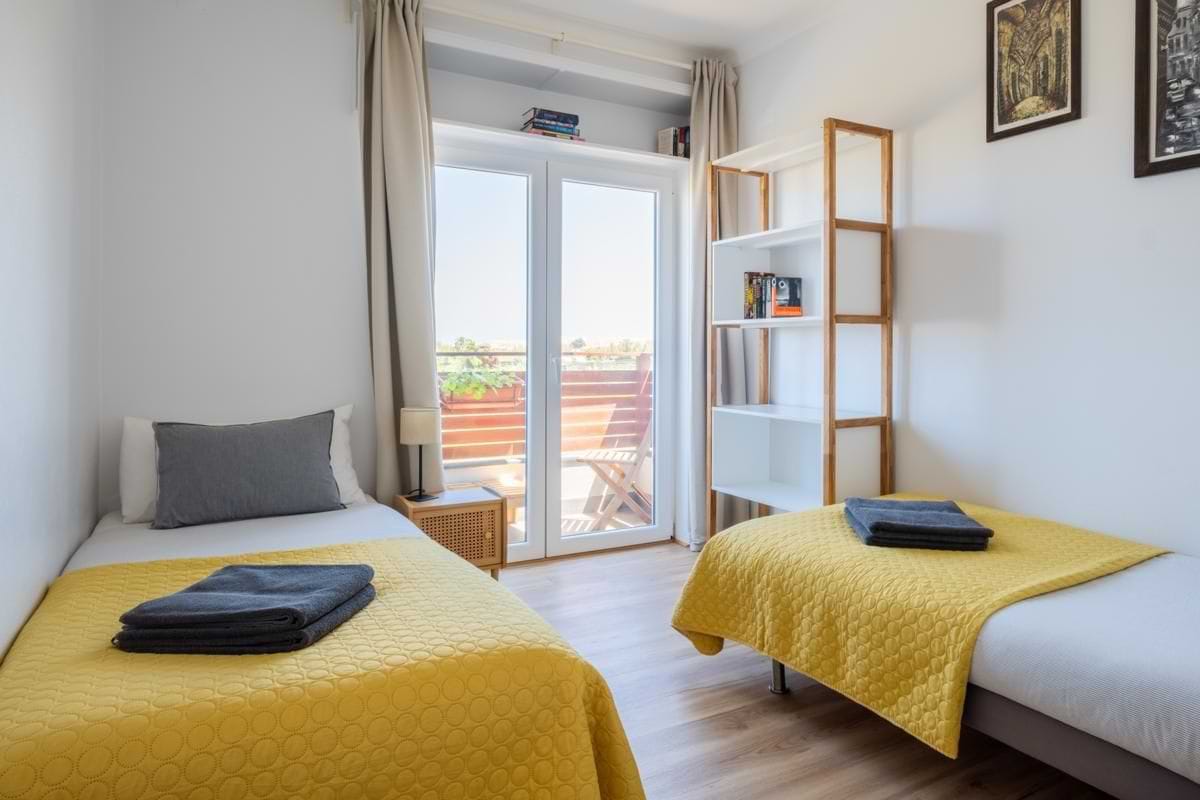 A bright bedroom with two single beds, each made with a distinctive yellow quilted throw and grey pillow, positioned opposite a glass door leading to a private balcony.