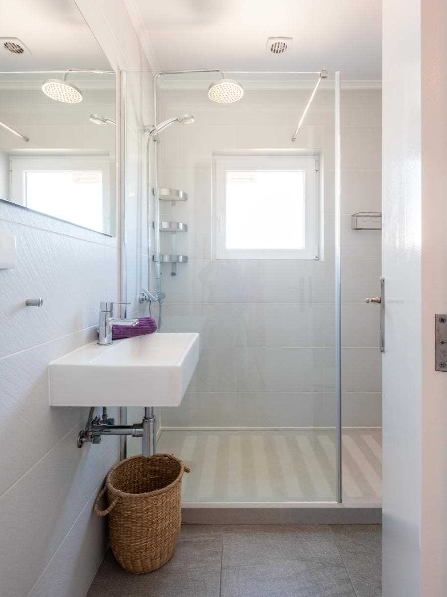 Bright white-tiled bathroom featuring a glass walk-in shower with a chrome rainfall head, a wall-hung sink, a mirror, and a small woven floor basket.