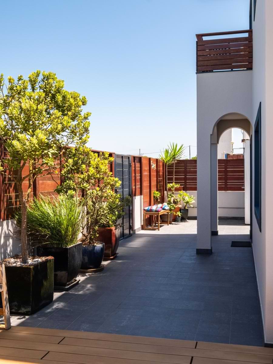 Tiled outdoor hallway and walkway lined with large potted trees and plants alongside a wooden privacy fence, leading toward the villa's arched doorway.